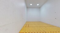 This single-level space is purpose-built for racquetball, offering a clean and well-maintained playing environment designed to meet the demands of the sport. The court features a smooth, light-toned hardwood floor finished with bold red regulation boundary and service lines, providing players with clear visual references during play. The floor's polished surface ensures consistent ball bounce and reliable footing throughout the court.

All four walls are finished in a bright white, smooth surface that reflects light evenly and allows for accurate ball rebound — a critical feature for competitive play. The walls show the characteristic markings typical of an actively used court, lending the space an authentic, well-used character.

Overhead, recessed square lighting panels are evenly distributed across the white ceiling, delivering uniform, shadow-free illumination across the entire playing surface. The ceiling height is generously proportioned, as required for regulation racquetball play, giving the space an open, unobstructed feel.

The overall ambiance of the court is clean, functional, and sport-focused. The combination of bright lighting, reflective white walls, and a warm hardwood floor creates an environment that is both visually crisp and practically optimized for high-performance racquetball play.