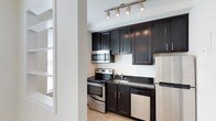 This single-floor space in Gallery, US, offers a clean and functional layout with a bright, open feel throughout. The main living area features light hardwood flooring and crisp white walls, creating an airy and welcoming atmosphere. A pass-through window connects the living space to an adjacent room, allowing natural light to flow freely and enhancing the sense of openness. The entry door and interior layout suggest a well-organized floor plan designed for comfortable everyday living.

The kitchen is a standout feature of the space, outfitted with rich dark cabinetry that contrasts elegantly against the light floors and walls. Dark granite countertops add a refined touch, while a full suite of stainless steel appliances — including a range, over-the-range microwave, dishwasher, and refrigerator — provides a modern, fully equipped cooking environment. Track lighting above the kitchen illuminates the workspace effectively, adding both function and a contemporary design element.

Overall, the space balances modern finishes with a practical layout, making it well-suited for a variety of uses. The consistent hardwood flooring, neutral color palette, and quality fixtures throughout contribute to a cohesive and polished aesthetic.