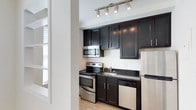 This single-floor space in Gallery, US, offers a clean and functional layout with a bright, open feel throughout. The main living area features light hardwood flooring and crisp white walls, creating an airy and welcoming atmosphere. A pass-through window connects the living space to an adjacent room, allowing natural light to flow freely and enhancing the sense of openness. The entry door and interior layout suggest a well-organized floor plan designed for comfortable everyday living.

The kitchen is a standout feature of the space, outfitted with rich dark cabinetry that contrasts elegantly against the light floors and walls. Dark granite countertops add a refined touch, while a full suite of stainless steel appliances — including a range, over-the-range microwave, dishwasher, and refrigerator — provides a modern, fully equipped cooking environment. Track lighting above the kitchen illuminates the workspace effectively, adding both function and a contemporary design element.

Overall, the space balances modern finishes with a practical layout, making it well-suited for a variety of uses. The consistent hardwood flooring, neutral color palette, and quality fixtures throughout contribute to a cohesive and polished aesthetic.