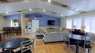 This single-floor space offers an open-concept layout that seamlessly integrates a living area, dining space, and kitchen into one cohesive environment. The vaulted ceiling with exposed beams adds architectural character and a sense of openness, while recessed lighting throughout ensures a bright and well-lit atmosphere.

The living area is furnished with comfortable gray sofas accented with colorful throw pillows, arranged around a central coffee table and anchored by a wall-mounted flat-screen television set against a bold periwinkle blue accent wall. This striking color choice adds a lively, welcoming energy to the space.

Adjacent to the living area, a dining section features sleek black tables paired with modern metal chairs, offering a practical and stylish setting for gatherings. The warm-toned hardwood flooring flows consistently throughout, tying the spaces together cohesively.

The kitchen, visible from the main living area, is outfitted with wood-toned cabinetry, a white range, microwave, and ample counter space, making it well-suited for meal preparation. Natural light filters in through windows along the exterior walls, complementing the recessed lighting above.

The overall ambiance is casual yet functional, designed to accommodate both social gatherings and everyday use with ease.