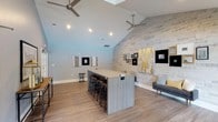 Located at 1500 Sunbow Falls Ln in Raleigh, NC, this single-floor space delivers a contemporary and welcoming atmosphere designed for gathering and relaxation. The room is anchored by soaring vaulted ceilings that create an expansive, airy feel, complemented by dual modern ceiling fans that ensure comfort throughout. A standout whitewashed brick accent wall runs the length of one side of the room, adding texture and character while serving as a backdrop for a curated collection of framed artwork in neutral and gold tones. Wide-plank hardwood-style flooring flows throughout, grounding the space with warmth and continuity. At the center, a generously sized island with a sleek laminate finish is surrounded by industrial-style barstools, making it ideal for casual seating or collaborative work. Recessed lighting is strategically placed throughout the ceiling to provide even, well-distributed illumination. A modern sofa and accent chair are arranged near the brick wall, offering a comfortable lounge area. The overall design blends industrial and contemporary elements seamlessly, resulting in a versatile, stylish space that is both functional and visually engaging.