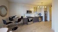 This contemporary single-floor residence offers a thoughtfully designed layout that balances comfort and modern style. The open-concept kitchen is outfitted with a full suite of stainless steel appliances, including a range, microwave, dishwasher, and refrigerator. Two-toned cabinetry — dark wood base cabinets paired with lighter upper cabinets — frames a white quartz countertop and a subway tile backsplash. A compact dining area with a bistro-style table sits adjacent, illuminated by a sleek track lighting fixture overhead. Light hardwood flooring runs throughout, lending warmth and continuity across the space. The living area is bright and welcoming, anchored by a tufted blue sofa, a glass coffee table, and a bold abstract artwork. A ceiling fan with integrated lighting adds function, while large windows fitted with horizontal blinds and a glass door provide natural light and views of the surrounding urban exterior. A sliding barn door leads to the bedroom, which is a serene retreat centered around a king-size bed beneath vibrant wall art. A walk-in closet offers ample storage. The bathroom is finished in crisp white tones, featuring a single undermount sink with chrome fixtures set into a dark wood vanity, a framed mirror with wall sconce lighting, and a full soaking tub with shower combination.