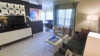 This thoughtfully designed single-floor residence offers an inviting open-concept layout that seamlessly connects the living and dining areas. The living room makes a striking first impression with a deep navy accent wall that anchors the space, providing a bold contrast against light neutral carpeting and crisp white trim details. A sleek media console and curated décor add a contemporary flair, while a plush sofa and a rounded accent chair create a comfortable and stylish seating arrangement around a modern chrome-framed coffee table. Large windows dressed with teal curtains and horizontal blinds flood the space with natural light and offer views of the greenery outside, enhancing the overall sense of openness. An elegant arched passageway leads into the dining area, where a round white table surrounded by upholstered chairs sits beneath warm ambient lighting. A vibrant artwork piece adds a pop of color to this intimate dining nook. The cohesive design language throughout — marked by a mix of modern furnishings, bold color choices, and carefully placed accents including fresh florals and decorative accessories — gives the space a polished, welcoming atmosphere suited for both everyday living and entertaining.