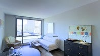This thoughtfully designed single-floor space offers a seamless blend of modern aesthetics and functional living. The living area is anchored by a large floor-to-ceiling window that frames an expansive urban skyline view, flooding the interior with abundant natural light throughout the day. The clean, neutral wall tones in soft white and grey create a calm, airy atmosphere that complements the contemporary furnishings.

The living area is furnished with a streamlined white sofa and coordinating seating, centered around a compact coffee table — an arrangement that maximizes both comfort and open floor space. A stylish dark-toned dresser with classic hardware sits against one wall, topped with a gold-accented mirror and task lamp, adding a touch of warmth and visual contrast to the otherwise light palette. Artwork displayed on the walls contributes a curated, intentional feel to the space.

The wood-toned flooring grounds the room with warmth while maintaining a sleek, modern look consistent throughout. The high, smooth ceiling enhances the sense of openness and volume. Overall, the space presents a polished, urban living environment defined by its connection to the city outside, its smart use of space, and its cohesive contemporary design language.
