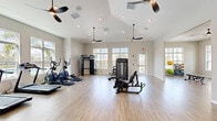 This generously sized fitness center offers a well-organized, single-floor layout designed to accommodate a full range of exercise activities. The space is defined by its vaulted ceilings, which rise prominently in the central area, creating an open, airy atmosphere enhanced by multiple ceiling fans and recessed lighting throughout.

Natural light is a defining feature, with large multi-pane windows lining the walls on multiple sides and glass-panel doors providing views of the surrounding outdoor environment. The bright, neutral gray walls and light wood-look flooring contribute to a clean, modern aesthetic that feels both welcoming and functional.

The equipment area is well-appointed, featuring treadmills, elliptical trainers, a multi-station cable machine, a leg press machine, a stationary bike, and a weight bench, among other strength and conditioning equipment. A wall-mounted flat-screen television is positioned for visibility across the space. A separate open area with high ceilings provides ample room for stretching or floor-based exercises. Additional rooms visible through interior glass-panel doors suggest supplementary amenity spaces nearby. Overall, the fitness center presents a polished, professional environment suited for comprehensive daily workouts.