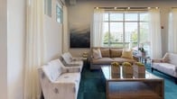 This sophisticated single-floor community amenity space is thoughtfully designed to offer residents a versatile gathering environment with a polished, contemporary aesthetic. The space is anchored by a spacious lounge area furnished with neutral-toned sofas and patterned accent chairs, set atop a rich teal carpet that ties together the refined color palette. Large floor-to-ceiling windows flood the interior with natural light and frame scenic outdoor views of a waterway and surrounding greenery, enhanced by sheer white drapery that adds an airy elegance. A bold abstract painting and textured wallcovering with wall sconces lend artistic character to the lounge nook. The dining area features multiple dark-topped tables with upholstered seating, positioned to take full advantage of the panoramic water views through expansive glazed walls. Soaring teal-painted ceilings and sleek track lighting accentuate the volume of this area. The demonstration kitchen is a standout feature, showcasing dark wood cabinetry, a white quartz countertop island with bar stool seating, stainless steel appliances, a gas range, built-in TVs, and light hardwood flooring — ideal for events and entertaining. Together, these zones create a cohesive, upscale communal environment.