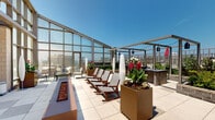 Located at 8621 Georgia Ave in Silver Spring, Maryland, this rooftop terrace offers a sophisticated indoor-outdoor amenity space with sweeping panoramic views of the surrounding cityscape. The space is defined by its striking floor-to-ceiling glass curtain wall pavilion, which floods the interior with abundant natural light and frames expansive views of the Silver Spring skyline on all sides.

The outdoor terrace is thoughtfully designed with large-format tile pavers and features multiple distinct seating areas. A row of wood-framed lounge chairs invites relaxation, while bistro-style tables and chairs offer a more intimate setting for gathering. Raised planter boxes filled with vibrant flowering plants and lush greenery add color and a garden-like ambiance throughout.

A linear fire feature runs along the terrace, providing a striking visual focal point and a warm gathering space. A pergola-style structure with pendant lighting defines an outdoor bar and lounge area, ideal for social events. An outdoor flat-screen television is mounted on the masonry accent wall, adding a functional entertainment element to the space.

Inside the glass-enclosed pavilion, comfortable lounge seating is positioned to take full advantage of the floor-to-ceiling city views, creating a bright, airy retreat. The combination of modern architectural details, curated landscaping, and unobstructed urban vistas makes this rooftop an exceptional communal destination.