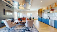 Located at 2225 Chester St in Aurora, CO, this stylish single-floor communal space blends bold contemporary design with functional comfort. The open-plan living room draws the eye upward to a striking sculptural ceiling installation featuring sweeping metallic curves and elegant pendant lanterns, creating a dramatic focal point overhead. Light hardwood floors in a herringbone pattern run throughout, complementing the bright, airy atmosphere generated by large windows on multiple walls that invite abundant natural light and open views of the surrounding neighborhood. A floor-to-ceiling textured tile accent wall anchors the living area, housing a sleek linear fireplace and a mounted display screen, flanked by vibrant large-format artwork that adds a creative, curated energy to the space. Seating areas are thoughtfully arranged with a mix of leather sofas, accent chairs, and intimate bistro tables, accommodating both relaxed lounging and casual gatherings. The kitchen features bold cobalt-blue cabinetry paired with gold hardware, a patterned tile backsplash, light countertops, and stainless steel appliances. Direct access to a rooftop outdoor terrace extends the space further, offering open-air seating, a grill station, and sweeping views of the Aurora skyline — ideal for entertaining.