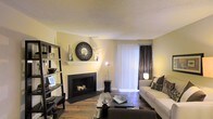 This single-floor residence offers a thoughtfully designed living space that balances comfort and style. The living room serves as the heart of the home, anchored by a sleek, black-framed fireplace topped with a decorative white mantel and an ornate circular mirror, adding a striking focal point to the room. Warm hardwood floors run throughout, lending a sense of richness and continuity to the space. Soft, neutral yellow-toned walls create a welcoming ambiance, complemented by natural light that filters in through floor-to-ceiling vertical blinds and elegant dark drape panels. A generously sized sofa and coordinating armchair are arranged around a dark glass coffee table, creating an ideal setting for relaxation or entertaining. A tall, open black shelving unit along one wall provides both display and storage space, adding a modern edge to the room's overall aesthetic. The textured ceiling adds a subtle architectural detail overhead. Together, these elements combine to create a living space that is both functional and visually appealing, offering a warm and inviting environment throughout.