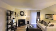 This single-floor residence offers a thoughtfully designed living space that balances comfort and style. The living room serves as the heart of the home, anchored by a sleek, black-framed fireplace topped with a decorative white mantel and an ornate circular mirror, adding a striking focal point to the room. Warm hardwood floors run throughout, lending a sense of richness and continuity to the space. Soft, neutral yellow-toned walls create a welcoming ambiance, complemented by natural light that filters in through floor-to-ceiling vertical blinds and elegant dark drape panels. A generously sized sofa and coordinating armchair are arranged around a dark glass coffee table, creating an ideal setting for relaxation or entertaining. A tall, open black shelving unit along one wall provides both display and storage space, adding a modern edge to the room's overall aesthetic. The textured ceiling adds a subtle architectural detail overhead. Together, these elements combine to create a living space that is both functional and visually appealing, offering a warm and inviting environment throughout.