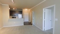 This single-floor residence offers a thoughtfully designed open-concept layout that seamlessly integrates the main living and kitchen areas. The kitchen is outfitted with dark espresso cabinetry, stainless steel appliances including a refrigerator, microwave, and range, and light-toned granite countertops that extend to a breakfast bar — ideal for casual dining or meal prep. Track lighting above the kitchen adds a modern, functional touch.

The living area flows naturally from the kitchen, featuring soft wall-to-wall carpeting that adds warmth and comfort underfoot, contrasting with the darker hard flooring in the kitchen and hallway. Crown molding runs along the ceiling throughout, lending an elevated, refined character to the space.

A central hallway connects the living area to the rest of the unit, with crisp white doors leading to additional rooms and closets. The clean, neutral color palette — warm beige walls paired with bright white trim — creates a welcoming and versatile backdrop. Flush ceiling lighting ensures consistent illumination throughout. Overall, the space strikes a balance between modern finishes and comfortable, practical living, making it a functional and inviting environment.