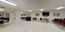 This single-floor community hall is designed to accommodate a variety of group activities and events, offering generous, open floor plans across several interconnected areas. The space features light-toned tile flooring and white walls throughout, creating a clean, bright atmosphere enhanced by recessed and overhead fluorescent panel lighting.

The main gathering area is expansive and flexible, furnished with rows of burgundy padded chairs that can be rearranged to suit different event formats. A classic upright piano anchors one wall, accompanied by a large wall-mounted flat-screen television and soundbar, making the space well-suited for presentations, performances, or community screenings. Colorful artwork and framed paintings are displayed throughout, adding warmth and character to the interior.

An adjoining room features long folding tables with matching burgundy chairs arranged for classroom or banquet-style seating, ideal for meetings, workshops, or dining. A prominent American flag and additional artwork decorate the walls, reinforcing the community-oriented nature of the space. Structural columns define zones within the open layout without interrupting the overall flow.

A service counter and water dispenser are available, supporting event hospitality needs. Emergency exit signage and a coat rack indicate thoughtful attention to functionality. The overall ambiance is welcoming, organized, and highly adaptable for civic, educational, or social gatherings.