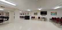 This single-floor community hall is designed to accommodate a variety of group activities and events, offering generous, open floor plans across several interconnected areas. The space features light-toned tile flooring and white walls throughout, creating a clean, bright atmosphere enhanced by recessed and overhead fluorescent panel lighting.

The main gathering area is expansive and flexible, furnished with rows of burgundy padded chairs that can be rearranged to suit different event formats. A classic upright piano anchors one wall, accompanied by a large wall-mounted flat-screen television and soundbar, making the space well-suited for presentations, performances, or community screenings. Colorful artwork and framed paintings are displayed throughout, adding warmth and character to the interior.

An adjoining room features long folding tables with matching burgundy chairs arranged for classroom or banquet-style seating, ideal for meetings, workshops, or dining. A prominent American flag and additional artwork decorate the walls, reinforcing the community-oriented nature of the space. Structural columns define zones within the open layout without interrupting the overall flow.

A service counter and water dispenser are available, supporting event hospitality needs. Emergency exit signage and a coat rack indicate thoughtful attention to functionality. The overall ambiance is welcoming, organized, and highly adaptable for civic, educational, or social gatherings.