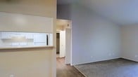 This single-floor residential space offers a clean, functional layout designed for comfortable everyday living. The interior is characterized by neutral wall tones in soft beige and cool gray, creating a calm and versatile backdrop throughout. The living area features warm wood-look hard flooring that transitions smoothly into plush carpeting, clearly delineating the distinct zones within the open floor plan. A defining architectural feature is the kitchen pass-through window, which connects the kitchen to the living area, promoting an open and connected feel. The kitchen is fitted with white cabinetry and appliances, maintaining a bright and cohesive aesthetic. A standout storage feature is the walk-in closet, visible through a doorway and offering generous storage capacity. The layout flows intuitively from one area to the next, with clear sightlines that enhance the sense of openness. Overall, the space presents a well-organized, move-in-ready environment with practical storage solutions, durable finishes, and a straightforward design that accommodates a variety of furnishing styles and living arrangements.