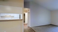 This single-floor residential space offers a clean, functional layout designed for comfortable everyday living. The interior is characterized by neutral wall tones in soft beige and cool gray, creating a calm and versatile backdrop throughout. The living area features warm wood-look hard flooring that transitions smoothly into plush carpeting, clearly delineating the distinct zones within the open floor plan. A defining architectural feature is the kitchen pass-through window, which connects the kitchen to the living area, promoting an open and connected feel. The kitchen is fitted with white cabinetry and appliances, maintaining a bright and cohesive aesthetic. A standout storage feature is the walk-in closet, visible through a doorway and offering generous storage capacity. The layout flows intuitively from one area to the next, with clear sightlines that enhance the sense of openness. Overall, the space presents a well-organized, move-in-ready environment with practical storage solutions, durable finishes, and a straightforward design that accommodates a variety of furnishing styles and living arrangements.