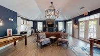 The Kensington's clubroom is a thoughtfully designed single-floor communal space that blends transitional style with inviting functionality. The open layout is anchored by a striking floor-to-ceiling stacked stone fireplace, which serves as the room's focal point and supports a wall-mounted television above the hearth. Vaulted ceilings add a sense of volume throughout, while a dramatic drum chandelier with Edison bulbs illuminates the central lounge area.

Deep navy accent walls create a bold, sophisticated backdrop that contrasts elegantly with warm hardwood-style flooring and crisp white wainscoting along the lower walls. The lounge is furnished with a rich cognac leather sofa, navy accent chairs, and layered area rugs, fostering a relaxed yet refined atmosphere.

The kitchenette features white shaker cabinetry, granite countertops, a white subway tile backsplash, a stainless steel sink, and built-in appliances, including a microwave and oven, accented by a rustic wood range hood shelf. A bistro-height dining table positioned near the windows and French doors provides a casual dining nook with views of the outdoors. A game area with a shuffleboard table rounds out the space, enhancing its recreational appeal.