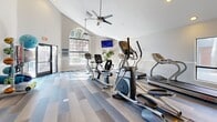 Located at 1055 W Baseline Rd in Mesa, Arizona, this fitness center offers a well-appointed exercise space designed to support a complete workout routine. The room features soaring vaulted ceilings that create an open, airy atmosphere, complemented by dual ceiling fans that ensure steady airflow throughout. Recessed lighting works alongside large arched windows to flood the space with natural light, enhancing the bright and motivating environment.

The flooring consists of stylish multi-tone plank in varying shades of gray and beige, providing both durability and a modern aesthetic. Cardio equipment is prominently featured, including treadmills, ellipticals, upright and recumbent stationary bikes, all from Star Trac. A functional trainer cable machine anchors the strength training section, while a well-organized rack holds a variety of medicine balls, resistance bands, yoga mats, and dumbbells for versatile workouts.

A wall-mounted flat-screen television adds entertainment during exercise sessions, and a classic wall clock keeps users on schedule. Decorative indoor plants are positioned throughout, softening the space and adding a touch of greenery. The layout is thoughtfully arranged to allow ample room for movement between stations, making this fitness center both functional and inviting for daily use.