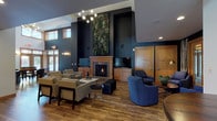 This thoughtfully designed single-floor community space blends modern sophistication with warm, inviting elements throughout. The centerpiece of the main lounge is a striking floor-to-ceiling living green wall flanking a wood-trimmed fireplace, creating a bold focal point against deep navy-painted walls. Soaring ceilings amplify the sense of openness, while a mid-century modern sputnik chandelier adds a refined touch overhead. Rich hardwood flooring runs throughout, tying together the various zones seamlessly.

Multiple seating arrangements offer flexible gathering options — light grey upholstered chairs and sofas anchor a central conversation area, while a cluster of deep blue swivel chairs near the fireplace provides a cozier nook. A wall-mounted television and built-in cabinetry enhance the entertainment functionality of the space.

A dedicated meeting or dining area features dramatic double-height windows with wood framing, flooding the room with natural light and offering views to the outdoors. Dark metal communal tables paired with industrial-style chairs complete this versatile zone.

The full kitchen is outfitted with warm wood cabinetry, granite countertops, black appliances, and a peninsula with leather bar stools — ideal for casual dining or entertaining. Together, these spaces form a well-appointed, multi-functional community environment.