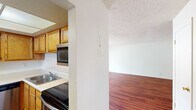 The Summit West Two Bedroom B1 unit offers a well-organized, single-floor layout designed for comfortable everyday living. The kitchen is efficiently arranged in an L-shape, featuring warm honey-toned oak cabinetry, stainless steel appliances including a side-by-side refrigerator with ice maker, dishwasher, microwave, and electric range, all set against light countertops and wood-look flooring. A bright drop-panel ceiling light fixture illuminates the space generously. Adjacent to the kitchen, the open living area is spacious and inviting, showcasing rich wood-look flooring, a textured ceiling, a ceiling fan, and a sliding glass door that leads to a covered patio. The patio, enclosed by white cinder block walls, offers a private outdoor retreat with views of surrounding greenery. The two bedrooms are well-proportioned, each with wood-look flooring, textured ceilings, and windows that bring in natural light. The primary bathroom includes a dual-sink vanity with ample counter space and mirrored medicine cabinets, while the second bathroom features a single-sink vanity and a full tub-shower combo with white tile surround. Throughout, the unit maintains a clean, neutral palette that provides a versatile and functional living environment.