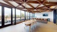 This architecturally distinctive single-floor space makes an immediate impression with its dramatic geometric wood-beam ceiling — a bold lattice of rich walnut-toned timbers forming a triangulated pattern that spans the entire length of the interior. Floor-to-ceiling windows line the perimeter, flooding the space with abundant natural light and offering expansive views of the surrounding treetops and cityscape beyond.

The open layout is thoughtfully organized into several functional zones. A central dining and meeting area features a large oval wood table surrounded by contemporary white and natural-toned chairs, ideal for gatherings or collaborative work. Adjacent to it, a fully appointed kitchenette includes sleek wood cabinetry, a white stone countertop, an undermount sink, stainless steel beverage coolers, and a wall-mounted television framed by textured white tile — combining practicality with refined aesthetics.

A lounge area anchors one end of the space, furnished with a curved teal velvet sofa, cognac leather swivel chairs, and soft accent pieces arranged around a linear fireplace set into a smooth white wall. Lush potted plants add warmth and a connection to nature throughout.

A glazed interior partition separates an additional flexible open area with patterned carpet flooring, suited for varied uses. Direct access to an outdoor terrace further enhances the space's appeal, seamlessly blending interior comfort with outdoor enjoyment.