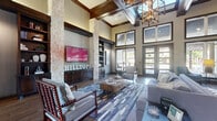 Located at 2200 N Loop 336 W in Conroe, Texas, this single-floor clubhouse at Hilltops offers a sophisticated and welcoming community gathering space. The living room commands attention with dramatic soaring ceilings adorned by a coffered wood-trimmed design, a geometric chandelier, and floor-to-ceiling windows that flood the interior with natural light and offer views of the surrounding outdoors. Flanking a large flat-screen television, natural stone columns and dark wood built-in shelving create a striking focal point. Rich hardwood flooring flows seamlessly throughout the entire space. A floor-to-ceiling natural stone fireplace with a rustic wood mantel anchors a secondary lounge area, accessible through elegantly arched dark wood doorways. The kitchen area showcases a standout curved island with a bold cobalt blue base and white stone countertop, illuminated by sleek pendant lighting, accompanied by tufted bar seating and stainless steel appliances. A dedicated game room features a billiards table and casual café-style seating, while a Starbucks coffee station provides a convenient amenity for residents. Together, these spaces deliver a refined yet comfortable ambiance ideal for socializing and relaxation.