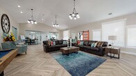This thoughtfully designed single-floor space blends modern farmhouse aesthetics with functional versatility. The most striking architectural feature is the soaring vaulted ceiling, which creates an expansive, airy atmosphere throughout the open-plan layout. Underfoot, a beautifully laid herringbone-patterned wood-look tile floor runs seamlessly across the entire space, adding warmth and visual texture.

The main gathering area is anchored by a pair of rich dark leather sofas arranged around a rustic wood coffee table, set atop a vibrant teal area rug. Pendant chandeliers with globe bulbs hang overhead, providing both ambient and statement lighting. A striking coral-red sliding barn door adds a bold pop of color and serves as a charming architectural accent.

Adjacent to the seating area, a stone-clad fireplace with a mounted television above it creates a cozy focal point, framed by large windows that invite natural light. A full-size pool table occupies its own dedicated zone, ideal for recreation and entertainment.

A glass-walled enclosure serves as a private office or conference area, offering transparency while maintaining separation from the main floor. A teal credenza with ornate detailing and decorative wall art complete the eclectic yet cohesive design. The overall ambiance is welcoming, modern, and rich in character.