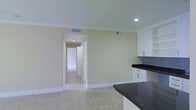 This single-floor space presents a clean, understated design characterized by light cream-colored walls, white crown molding, and neutral tile flooring throughout. The kitchen area is equipped with white cabinetry featuring simple bar-pull hardware and striking dark granite countertops that provide a bold contrast against the lighter surroundings. Recessed lighting illuminates the space efficiently, complemented by ceiling-mounted HVAC vents that contribute to a tidy, streamlined ceiling plane. A wide open doorway connects the main area to a hallway, which leads further into additional rooms of the unit, suggesting a functional and straightforward layout. The hallway benefits from its own recessed lighting, maintaining consistent brightness throughout. The overall ambiance is minimal and move-in ready, with a neutral color palette that offers versatility for a variety of interior design styles. The open sightlines between the kitchen and the connecting corridor enhance the sense of flow, making the space feel open and easy to navigate.
