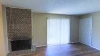 This single-floor space offers a warm and functional layout centered around a standout living area. The room is anchored by a classic full-brick fireplace, adding a timeless architectural element and a cozy focal point to the space. Rich hardwood flooring runs throughout, providing a durable and visually appealing foundation that complements the neutral, earthy wall tones.

A large sliding glass door fitted with vertical blinds serves as both a source of natural light and a direct connection to the outdoors. When the blinds are open, light floods the interior, enhancing the open and airy feel of the room. The textured popcorn ceiling adds a layer of acoustic and visual interest overhead.

The neutral color palette of warm beige walls paired with the deep tones of the hardwood floors and the rustic brick of the fireplace create a cohesive and inviting atmosphere throughout. The overall design lends itself well to a variety of furnishing styles, from traditional to contemporary. The space strikes a balance between classic architectural character and everyday practicality, making it a comfortable and versatile environment suited for relaxed living.