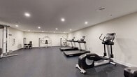 Located at 2-48 Grove Ave, this fitness center offers a comprehensive exercise facility designed to accommodate a wide range of workout routines. The space is thoughtfully laid out across a single floor, providing ample room for both cardio and strength training activities without feeling crowded.

The interior features clean white walls, a smooth, neutral ceiling with recessed lighting that ensures bright, even illumination throughout. The durable rubber flooring in a dark speckled finish provides cushioning and traction, making it well-suited for high-impact workouts.

Cardio equipment is well-represented, with multiple Life Fitness treadmills and elliptical cross-trainers positioned to give users sufficient personal space. For strength training, the facility includes a cable machine system, several selectorized weight machines, and dedicated free weight stations stocked with a variety of dumbbells neatly arranged on racks. Colorful resistance tools and exercise accessories add further variety to available workouts.

A wall-mounted flat-screen television and a clock are visible, contributing to a functional and comfortable environment. A mirrored wall section enhances the sense of openness and supports form-conscious training. The facility is accessed through a standard door and maintains a clean, organized atmosphere that supports focused, effective exercise sessions.