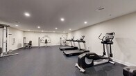 Located at 2-48 Grove Ave, this fitness center offers a comprehensive exercise facility designed to accommodate a wide range of workout routines. The space is thoughtfully laid out across a single floor, providing ample room for both cardio and strength training activities without feeling crowded.

The interior features clean white walls, a smooth, neutral ceiling with recessed lighting that ensures bright, even illumination throughout. The durable rubber flooring in a dark speckled finish provides cushioning and traction, making it well-suited for high-impact workouts.

Cardio equipment is well-represented, with multiple Life Fitness treadmills and elliptical cross-trainers positioned to give users sufficient personal space. For strength training, the facility includes a cable machine system, several selectorized weight machines, and dedicated free weight stations stocked with a variety of dumbbells neatly arranged on racks. Colorful resistance tools and exercise accessories add further variety to available workouts.

A wall-mounted flat-screen television and a clock are visible, contributing to a functional and comfortable environment. A mirrored wall section enhances the sense of openness and supports form-conscious training. The facility is accessed through a standard door and maintains a clean, organized atmosphere that supports focused, effective exercise sessions.