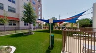 This thoughtfully designed outdoor pet area serves as a dedicated amenity space within a modern, multi-story residential community. The grounds are covered in lush, well-maintained artificial turf that provides a clean and comfortable surface throughout. Decorative landscaped planters with ornamental grasses and young trees are arranged throughout the green space, adding natural visual interest and a park-like atmosphere.

The area is divided into distinct functional zones, including a generously sized open turf lawn ideal for free roaming, and a separate paved enclosure featuring pet water stations and waste disposal units for added convenience. Colorful triangular shade sails in blue and red are suspended overhead in the paved section, offering relief from direct sunlight while string lights are strung above, lending a welcoming ambiance during evening hours.

The surrounding architecture features a combination of stone and brick facades with arched entryways, terra cotta accent rooflines, and multi-level balconies, reflecting a Mediterranean-inspired design aesthetic. Iron fencing with a dark bronze finish neatly defines the perimeter of the space, ensuring a secure and enclosed environment. The overall setting is bright, open, and well-organized, offering a clean and functional outdoor retreat within the larger residential complex.