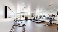 Located at 12444 Albrook Dr in Denver, Colorado, this well-equipped fitness center offers a comprehensive exercise environment designed to support a wide range of workout routines. The space features a clean, modern aesthetic with white walls, recessed lighting, and durable rubberized flooring that provides both comfort and functionality underfoot.

The room is thoughtfully organized to maximize its footprint. Cardio equipment — including multiple treadmills, an elliptical, and a stationary bike — lines one side of the space, complemented by a large wall mirror that visually expands the room and assists with form during exercise. A rowing machine and adjustable weight bench add versatility for strength and endurance training, while a full dumbbell rack offers a wide range of free weight options. TRX suspension training straps are mounted to the wall, enabling bodyweight resistance exercises. Medicine balls and additional functional fitness accessories round out the equipment selection.

Large windows along one wall flood the space with natural light and provide views of the surrounding outdoor area, creating an open and energizing atmosphere. Motivational artwork depicting runners is displayed prominently, adding a dynamic visual element to the environment. The space also benefits from climate control via ceiling vents and offers convenient access through multiple doorways. Overall, this fitness center is a practical, well-appointed facility suited for diverse training needs.