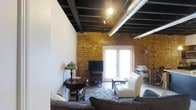 This single-floor loft space makes a striking architectural statement through its seamless fusion of industrial and contemporary design elements. The most commanding feature is the exposed brick wall, rendered in warm amber and ochre tones, which lends the space a sense of history and tactile richness. Overhead, dark-painted structural beams and exposed silver ductwork run the length of the ceiling, reinforcing the loft's industrial character while adding considerable visual height and drama.

Track lighting mounted along the ceiling beams provides focused illumination throughout the space, complementing the natural light that pours in through a set of elegant French doors. These glass-paned doors serve as both a bright focal point and a gateway to the outside, flooding the interior with an airy brightness that softens the rawness of the industrial elements.

The living area is thoughtfully appointed with a plush sectional sofa, a classic armchair in cream upholstery, and a patterned area rug anchoring the seating arrangement. A vintage side table with a Tiffany-style lamp adds a warm, eclectic touch, while a bar cart and entertainment setup complete the room's functional yet stylish layout. The overall ambiance is one of urban sophistication — a space that celebrates architectural authenticity.