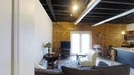 This single-floor loft space makes a striking architectural statement through its seamless fusion of industrial and contemporary design elements. The most commanding feature is the exposed brick wall, rendered in warm amber and ochre tones, which lends the space a sense of history and tactile richness. Overhead, dark-painted structural beams and exposed silver ductwork run the length of the ceiling, reinforcing the loft's industrial character while adding considerable visual height and drama.

Track lighting mounted along the ceiling beams provides focused illumination throughout the space, complementing the natural light that pours in through a set of elegant French doors. These glass-paned doors serve as both a bright focal point and a gateway to the outside, flooding the interior with an airy brightness that softens the rawness of the industrial elements.

The living area is thoughtfully appointed with a plush sectional sofa, a classic armchair in cream upholstery, and a patterned area rug anchoring the seating arrangement. A vintage side table with a Tiffany-style lamp adds a warm, eclectic touch, while a bar cart and entertainment setup complete the room's functional yet stylish layout. The overall ambiance is one of urban sophistication — a space that celebrates architectural authenticity.