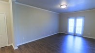 This single-floor space presents a clean, well-maintained interior highlighted by rich, dark hardwood-style flooring that runs throughout the main living area. The floors offer a striking contrast against the crisp white baseboards and crown molding that elegantly frame the room, adding a polished, finished look to the overall design.

The walls are painted in a soft, neutral tone that creates a calm and versatile backdrop, suitable for a variety of décor styles. Crown molding along the ceiling perimeter adds a subtle architectural detail that elevates the space beyond standard construction. A flush-mount ceiling light fixture provides overhead illumination, complementing the natural light that filters in through a windowed exterior door fitted with sheer curtains on the far wall.

The layout feels open and unobstructed, offering flexibility in how the space can be arranged and utilized. The windowed door suggests access to an outdoor area, allowing for natural light to enhance the room's airy feel. Overall, the space combines practical finishes with tasteful detailing, resulting in a bright, welcoming environment ready for immediate use.