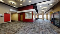 This single-floor common area is designed to serve as a welcoming hub for residents, combining functional services with comfortable social spaces. The main lobby greets visitors with a spacious, open layout accented by bold crimson and warm tan walls, patterned multi-tone carpet tiles, and recessed circular ceiling lighting. A glassed-in reception or management office provides an on-site staffing station, while a bank of resident mailboxes and a dedicated elevator lobby with two elevators and stairwell access ensure smooth building circulation. The game room is a standout amenity, featuring a full-size pool table with overhead television, swivel lounge chairs, and large windows that flood the space with natural light and views of the surrounding streetscape. Adjacent to this is a cozy seating lounge with teal upholstered sofas, patterned cushions, and oversized windows overlooking a landscaped outdoor area with mature trees. A community kitchen is equipped with warm wood cabinetry, a stainless steel sink, solid-surface countertops, and a dishwasher. Vending machines are conveniently located near the elevator core. Throughout, structural columns, black-accented ceiling soffits, and consistent flooring tie the various zones together into a cohesive, modern aesthetic well-suited for a residential community.