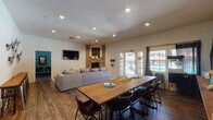 Located at 4601 E Skyline Dr in Tucson, Arizona, this single-story clubhouse offers a well-appointed gathering space designed for both relaxation and recreation. The main living area is anchored by a striking corner brick fireplace, adding a warm, rustic element to the otherwise contemporary interior. A large sectional sofa faces a wall-mounted flat-screen TV, creating an inviting lounge area ideal for group gatherings. Recessed lighting throughout ensures the space is well-lit, while wood-look flooring adds a cohesive, modern aesthetic. Adjacent to the living area, a generously sized wooden dining table with seating for numerous guests provides an excellent space for communal dining or meetings. Large French doors and multiple windows along the exterior wall flood the space with natural light and offer direct views of an outdoor pool area. A separate game room, accessible through double doors adorned with a dramatic full-wall mural of the Tucson desert landscape, features a full-size pool table set against teal-painted walls, carpeted flooring, and framed southwestern artwork. The overall ambiance of the space blends casual comfort with recreational functionality, reflecting the character of its Tucson surroundings.