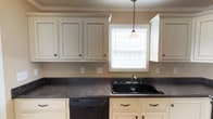 This two-story home offers a thoughtfully designed layout that balances functionality and comfort across both floors. The kitchen stands out as a well-equipped space, featuring cream-painted cabinetry with dark hardware accents and crown molding details that lend a timeless, traditional character. Dark laminate countertops provide a striking contrast against the light cabinetry, while a deep black drop-in sink sits beneath a window dressed with horizontal blinds, allowing natural light to filter into the space. A dishwasher is seamlessly integrated into the cabinetry for added convenience, and a pendant light hangs overhead, contributing warm, focused illumination. The warm beige wall tones throughout create a neutral and welcoming atmosphere that complements the classic design elements. Crown molding details visible along the ceiling add an elevated touch to the interior. The two-floor layout provides ample space for living and accommodating various needs, with each level contributing to the overall sense of a well-structured and practical home. The overall ambiance is cozy and traditional, with careful attention to detail evident in the finishes and fixtures throughout the space.
