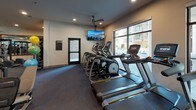 Located at 5619 Belmont Ave in Dallas, TX, this modern fitness center is thoughtfully designed to support a full range of workout routines. The single-floor space is laid out to maximize usability, with distinct zones for cardio and strength training. The cardio area features a row of Matrix treadmills and elliptical machines positioned alongside large windows that invite natural light and offer views of the surrounding outdoor area, creating an open and energizing atmosphere. A wall-mounted flat-screen TV adds to the experience for those logging miles on the treadmill. The strength training section is impressively outfitted with a Jones power rack, cable machines, selectorized resistance equipment, and a collection of free weights including dumbbells and plate-loaded barbells. Floor-to-ceiling mirrors throughout the weight area visually expand the space and provide functional feedback during lifts. A dedicated functional training zone offers stability balls and accessories for dynamic workouts. The flooring transitions between patterned blue carpet and rubberized mat surfaces appropriate for each zone. Ceiling fans and recessed lighting maintain comfort throughout. A geometric-tiled accent wall adds a contemporary design touch, while built-in storage cubbies and a compact beverage refrigerator offer added convenience. The overall ambiance is sleek, clean, and professional.