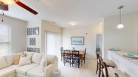 This single-floor residence is thoughtfully designed with a cohesive, light-filled aesthetic throughout. The open-concept living and dining area forms the heart of the unit, where plush carpet flooring, neutral beige walls, and a ceiling fan create a comfortable, airy atmosphere. A large sectional sofa anchors the living space, complemented by natural light streaming through wide windows fitted with horizontal blinds. A sliding glass door provides access to the exterior, further enhancing the brightness of the space.

The dining area flows seamlessly from the living room and features a wooden dining table with seating for multiple guests, accented by a coastal-themed framed artwork on the wall. The kitchen is well-appointed with dark espresso cabinetry, granite-style countertops, hardwood-style flooring, black appliances, and a double pantry with paneled doors, offering ample storage and functionality.

The bedrooms are carpeted and receive natural light through standard windows. The primary bathroom includes dark vanity cabinetry with granite countertops, a large mirror, and a shower with curtain. A second bathroom features a soaking tub with a shower curtain and matching dark cabinetry, maintaining the unit's consistent design language throughout.