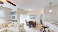 This single-floor residence is thoughtfully designed with a cohesive, light-filled aesthetic throughout. The open-concept living and dining area forms the heart of the unit, where plush carpet flooring, neutral beige walls, and a ceiling fan create a comfortable, airy atmosphere. A large sectional sofa anchors the living space, complemented by natural light streaming through wide windows fitted with horizontal blinds. A sliding glass door provides access to the exterior, further enhancing the brightness of the space.

The dining area flows seamlessly from the living room and features a wooden dining table with seating for multiple guests, accented by a coastal-themed framed artwork on the wall. The kitchen is well-appointed with dark espresso cabinetry, granite-style countertops, hardwood-style flooring, black appliances, and a double pantry with paneled doors, offering ample storage and functionality.

The bedrooms are carpeted and receive natural light through standard windows. The primary bathroom includes dark vanity cabinetry with granite countertops, a large mirror, and a shower with curtain. A second bathroom features a soaking tub with a shower curtain and matching dark cabinetry, maintaining the unit's consistent design language throughout.