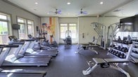 This fitness center offers a comprehensive and well-organized workout environment designed to accommodate a wide range of exercise needs. The space is laid out across a single floor, maximizing functionality and ease of movement between equipment zones.

The cardio section features a row of treadmills and elliptical machines, providing ample options for aerobic training. Complementing these are dedicated strength training stations, including a multi-function cable machine and a weight bench, offering users a full-body workout experience.

The flooring consists of durable rubber matting, providing both safety and comfort during exercise. Two ceiling fans circulate air throughout the room, helping to maintain a comfortable atmosphere during intense workouts. Recessed lighting ensures the space is well-illuminated at all times.

Large windows along the walls allow natural light to fill the room while offering views of the surrounding greenery, creating an energizing and uplifting environment. A wall-mounted mirror on one side enhances the sense of openness and allows users to monitor their form. A vibrant, colorful graphic mural adds a dynamic and motivating visual element to the space, contributing to its energetic ambiance. Overall, this fitness center is thoughtfully designed to support an active and healthy lifestyle.