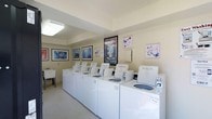 This shared laundry room is a practical and thoughtfully designed space that goes beyond basic functionality. A row of white top-loading washing machines lines the main wall, providing ample capacity for residents. The machines are well-maintained and clearly labeled for ease of use.

The room is finished with light-colored walls and tile flooring, contributing to a clean and bright environment. Overhead fluorescent lighting ensures the space is well-lit throughout. A utility sink is also available along one side wall, adding convenience for pre-treating garments or handwashing items.

What sets this laundry room apart is its attention to ambiance. The walls are decorated with an eclectic mix of framed artwork, including nature-inspired prints featuring floral motifs, serene landscape photography, and a vintage-style Café Martin poster, lending the space a welcoming and almost café-like character.

A notice board is mounted on the wall, providing a practical communication point for residents. Overall, the space balances utility with a pleasant aesthetic, making routine laundry tasks a more comfortable experience.