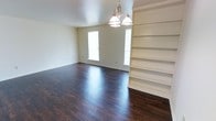 This single-floor residence presents a well-maintained and thoughtfully designed living space. The main living area immediately draws attention with its rich, dark hardwood flooring that runs throughout, providing a warm and polished aesthetic. Natural light flows in through multiple windows, brightening the neutral, light-toned walls and creating an open, airy atmosphere. A classic multi-bulb chandelier serves as a central lighting fixture, adding a traditional touch to the room's character. One of the standout architectural features is the built-in floor-to-ceiling shelving unit positioned along one wall, offering generous display and storage space while contributing to the room's refined, organized feel. The clean lines of the shelving complement the simplicity of the overall design. The space is finished with white baseboards that provide a crisp contrast against the dark flooring. The layout is open and unobstructed, allowing for flexible furniture arrangements and a sense of spaciousness. The overall ambiance is clean, calm, and move-in ready, with classic design elements that offer both practicality and visual appeal throughout the home.