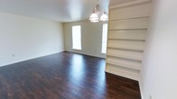 This single-floor residence presents a well-maintained and thoughtfully designed living space. The main living area immediately draws attention with its rich, dark hardwood flooring that runs throughout, providing a warm and polished aesthetic. Natural light flows in through multiple windows, brightening the neutral, light-toned walls and creating an open, airy atmosphere. A classic multi-bulb chandelier serves as a central lighting fixture, adding a traditional touch to the room's character. One of the standout architectural features is the built-in floor-to-ceiling shelving unit positioned along one wall, offering generous display and storage space while contributing to the room's refined, organized feel. The clean lines of the shelving complement the simplicity of the overall design. The space is finished with white baseboards that provide a crisp contrast against the dark flooring. The layout is open and unobstructed, allowing for flexible furniture arrangements and a sense of spaciousness. The overall ambiance is clean, calm, and move-in ready, with classic design elements that offer both practicality and visual appeal throughout the home.