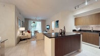 This single-floor model residence offers a thoughtfully designed open-concept layout that seamlessly connects the kitchen, dining, and living areas into one cohesive, light-filled space. The kitchen features rich wood cabinetry paired with striking dark granite countertops on a peninsula island, providing both generous prep space and a natural visual anchor to the room. Light-toned tile flooring runs throughout, lending a clean, cohesive aesthetic that enhances the sense of openness.

The living area is bathed in natural light, thanks to floor-to-ceiling sliding glass doors framed by sheer draping curtains that lead directly to a private outdoor balcony. A ceiling fan overhead keeps the space comfortable while adding a casual, relaxed touch. Neutral-toned furnishings, a plush area rug, and curated decorative accents — including teal artwork and metallic vases — complete the contemporary aesthetic.

The overall ambiance is bright, airy, and modern, with a clean color palette of whites and warm wood tones that creates an inviting and stylish atmosphere. The layout maximizes flow and functionality, making it an excellent representation of comfortable, contemporary living.