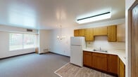 This single-floor unit in Grand Rapids, Michigan, offers a straightforward, functional layout with a seamless flow between the kitchen and living areas. The kitchen is well-equipped, featuring warm oak cabinetry that extends along the walls, providing generous storage space. A skylight-style fluorescent fixture brightens the cooking area, which includes a white refrigerator, an electric cooktop, a single-basin sink, and ample countertop workspace in an L-shaped configuration. Wood-look flooring grounds the kitchen zone, transitioning into carpeted living and dining areas.

The open dining and living space is illuminated by a brass chandelier and wall sconces, adding a classic touch to the otherwise neutral interior. A window in the living area allows natural light to filter in, offering a view of outdoor greenery. White walls and neutral carpet contribute to a clean, understated aesthetic throughout.

The bathroom is functionally appointed with a single-sink vanity topped with a light countertop, a large wall mirror with overhead lighting, and a tiled tub-shower combination in neutral beige tones. A closet with wood-paneled doors provides additional storage nearby. Overall, the space presents a practical and comfortable layout suitable for everyday living.