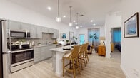 This single-floor residence showcases a thoughtfully designed open-concept layout that seamlessly connects the kitchen, dining, and living areas. The kitchen is a standout feature, outfitted with gray shaker-style cabinetry, granite countertops, a neutral tile backsplash, and a full suite of stainless steel appliances including a range, microwave, dishwasher, and refrigerator. A central island with granite countertop and bar seating serves as both a functional workspace and casual dining spot, illuminated overhead by a trio of globe pendant lights.

The living area is bright and welcoming, with light hardwood-style flooring that carries throughout the main living spaces. A glass-paneled door allows natural light to filter in and provides outdoor access. The space features elevated ceilings that contribute to an airy, open atmosphere.

The bathroom is clean and modern, featuring gray cabinetry with granite countertops, a glass-enclosed walk-in shower with tiled walls, and light flooring that ties into the overall aesthetic of the home. The bedroom offers a calm, neutral retreat with soft carpeting and ample wall space. Throughout, the design maintains a cohesive coastal-contemporary palette of whites, grays, and natural wood tones.