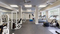 Located at 809 E Main St in Lexington, SC, this fitness center offers a well-equipped, single-floor layout designed to support a variety of exercise routines. The main workout area is generously sized, with structural columns defining distinct zones while maintaining an open, airy feel. Durable rubber flooring covers the entire exercise space, providing both safety and functionality for a range of activities.

The cardio section features multiple treadmills and a stair climber, positioned near large windows that allow natural light to fill the room and offer views of the outdoor pool area, creating an inviting atmosphere for exercise. Ceiling fans are distributed throughout the space, supplemented by overhead fluorescent lighting to ensure consistent illumination.

The strength training area is equipped with white cable weight machines and a weight bench with free weights, offering users a comprehensive resistance workout. Wall-mounted mirrors in the strength zone allow for form monitoring during exercises.

A separate, adjacent room serves as a dedicated children's play area, outfitted with colorful foam floor tiles and age-appropriate toys, making the facility family-friendly. The overall interior features neutral beige walls, white trim, and textured ceilings, contributing to a clean and functional aesthetic throughout the space.