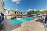 Located at 710 S College Rd in Lafayette, Louisiana, this outdoor pool area serves as a central amenity within a multi-building residential complex. The centerpiece is a generously sized rectangular swimming pool with vivid turquoise water, framed by a classic brick coping border and accessible via stainless steel entry rails. The pool deck is finished with large-format flagstone pavers in warm earth tones, creating a durable and visually appealing surface that wraps extensively around all sides of the pool. Lounge chairs are positioned throughout the deck, offering ample space for sunbathing and relaxation. Patio umbrellas provide shaded seating areas, enhancing the usability of the outdoor space during sunny Louisiana days. The pool area is fully enclosed by a black iron perimeter fence, providing security and clear boundaries. The surrounding residential buildings feature a distinctive architectural style, with slate-style shingle siding, wood-toned trim, and covered upper-level balconies connected by exterior staircases. Mature trees visible beyond the buildings contribute greenery and shade to the overall setting. The combination of the inviting pool, well-maintained stone patio, and the classic architectural backdrop creates a comfortable and functional outdoor communal space suited for warm-weather enjoyment.